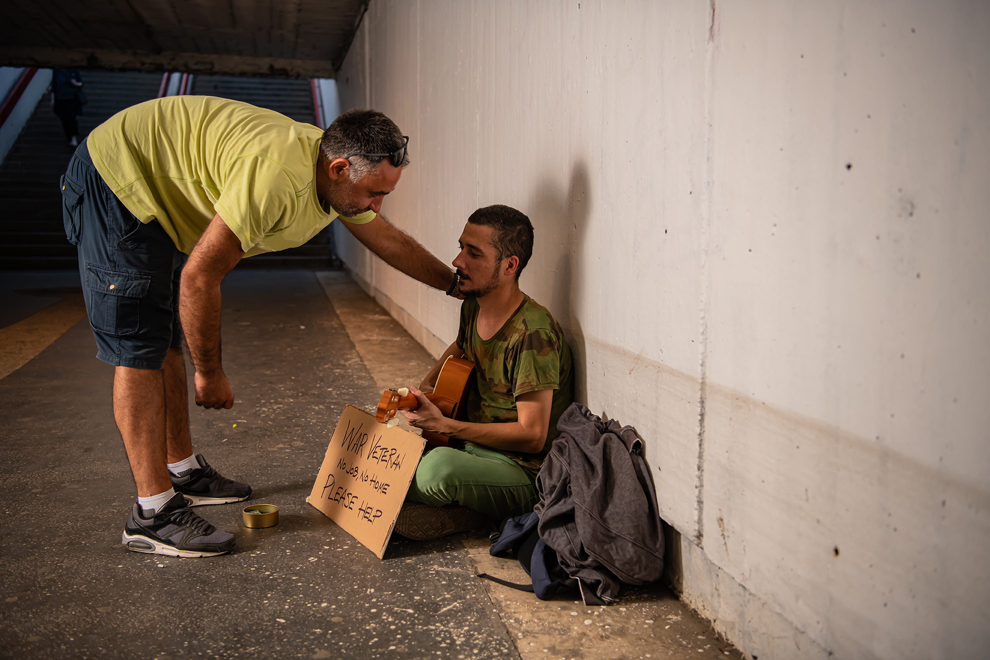 white man standing and bending down to put a hand on the shoulder of a young white man seated on the ground holding a sign that says "war veteran no job, no home please help" and playing guitar
